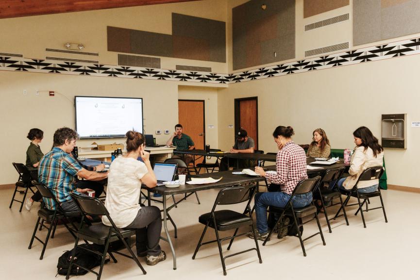 People sitting, conversing, and thinking at tables grouped in a circle. 