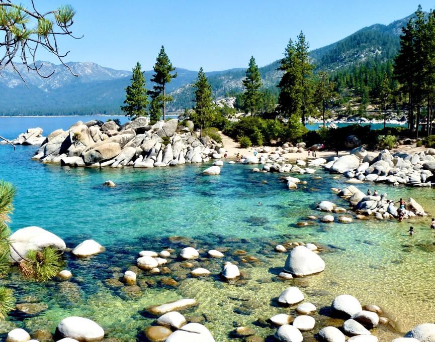 people and kids play on a sandy and bolder-filled beach near crystal clear lake waters