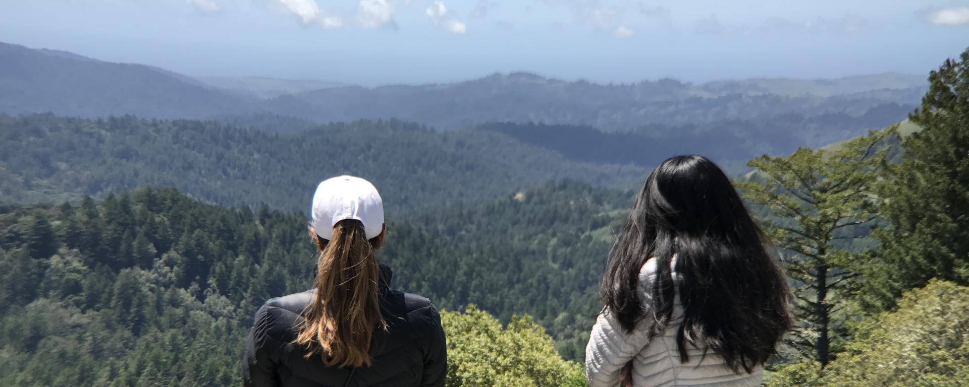 Two women stand on a hilltop looking out over a forested landscape