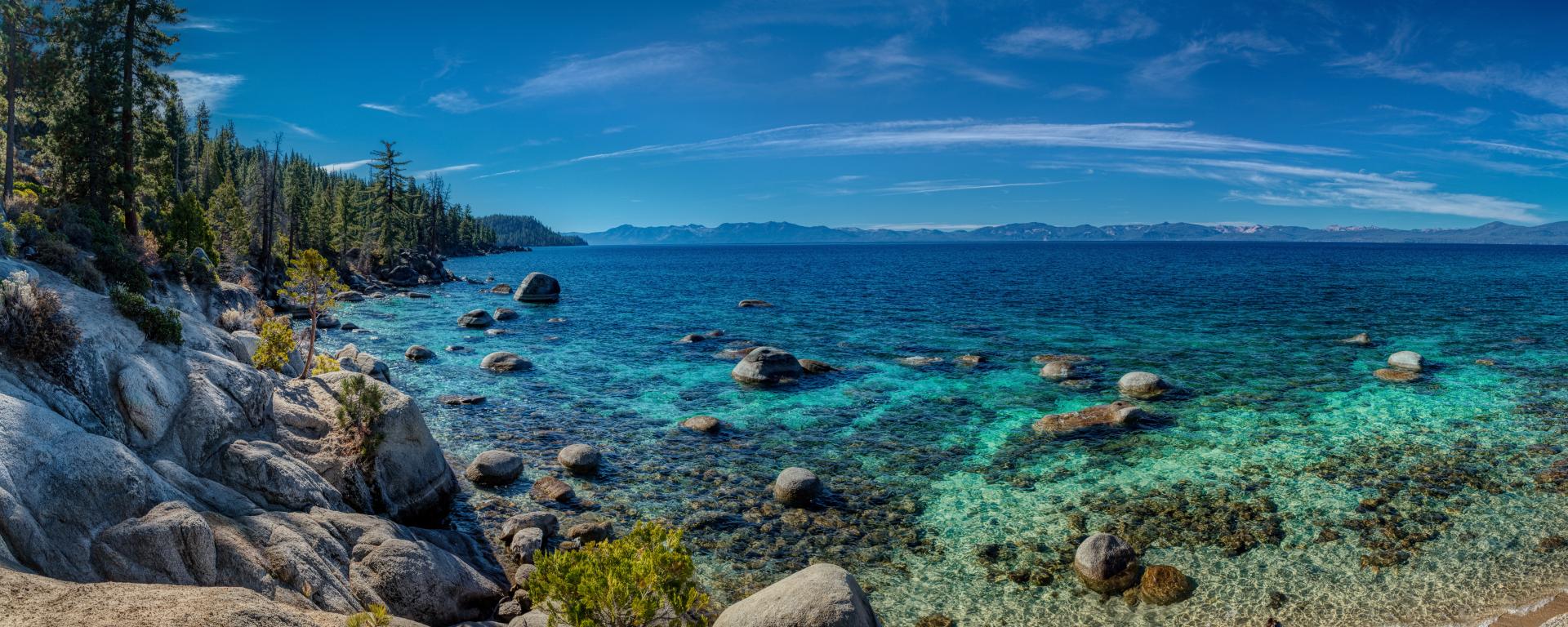 Looking out from a rocky, tree-lined coast at blue water and blue sky, with mountains lining the distant horizon.